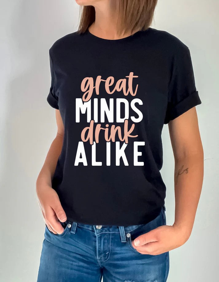 Woman wearing a black Gildan T-shirt with bold text reading “Great Minds Drink Alike” in white and tan lettering, paired with jeans.