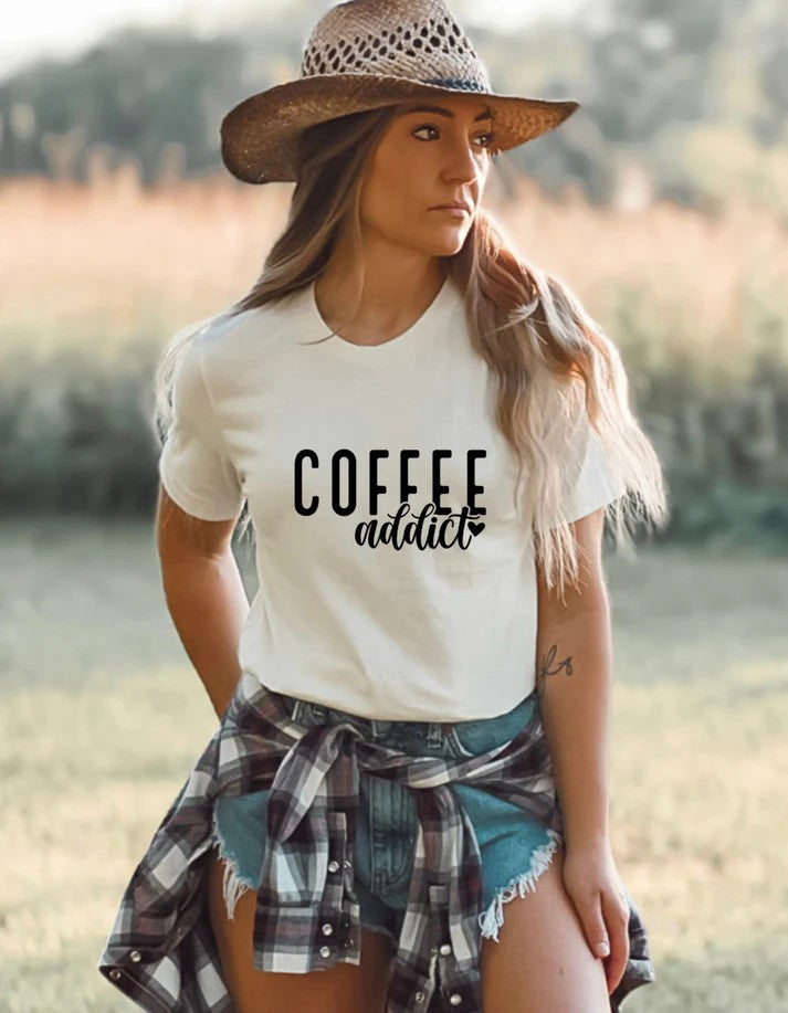 Woman wearing a white Gildan T-shirt with black text reading “Coffee Addict,” styled with denim shorts, a plaid shirt tied at the waist, and a straw hat in an outdoor setting.