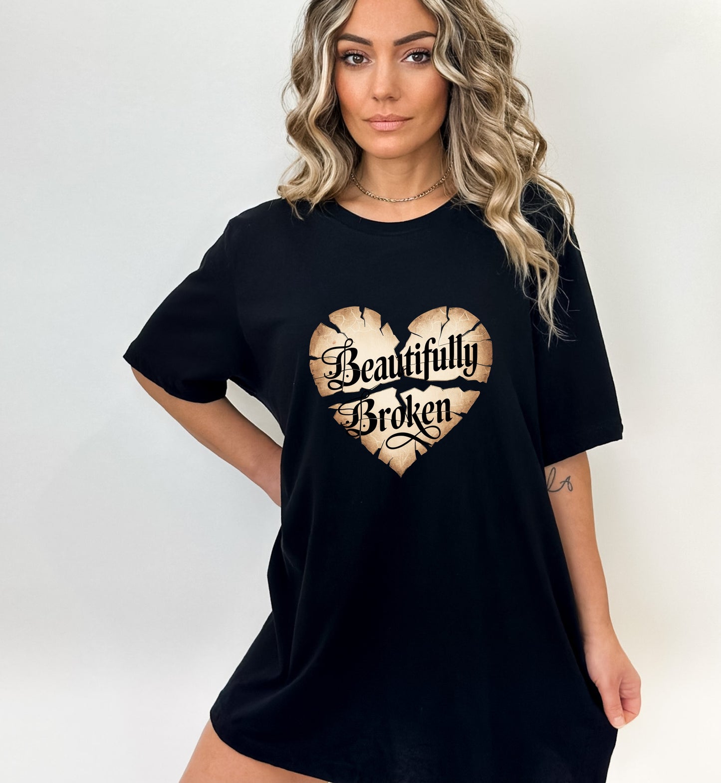 Woman wearing a black oversized Gildan T-shirt with a cracked beige heart design and the words “Beautifully Broken” in ornate black script on the front.