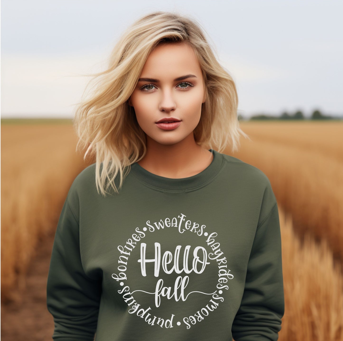 Blonde woman wearing an olive green sweatshirt with the phrase “Hello Fall” surrounded by words like bonfires, sweaters, hayrides, s’mores, and pumpkins in white script font, standing in a golden wheat field.