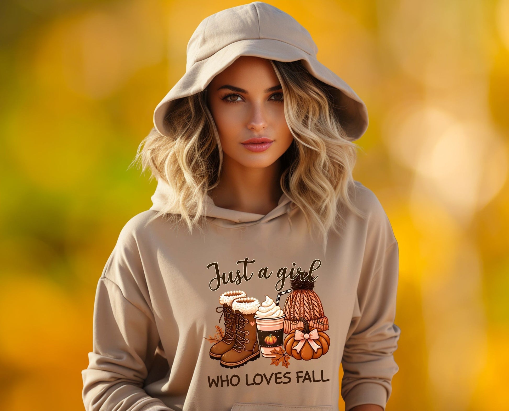 Blonde woman wearing a beige hoodie with the text “Just a Girl Who Loves Fall” and graphics of fall boots, pumpkin spice coffee, a knit hat, and pumpkins, standing in front of a blurred golden autumn background.