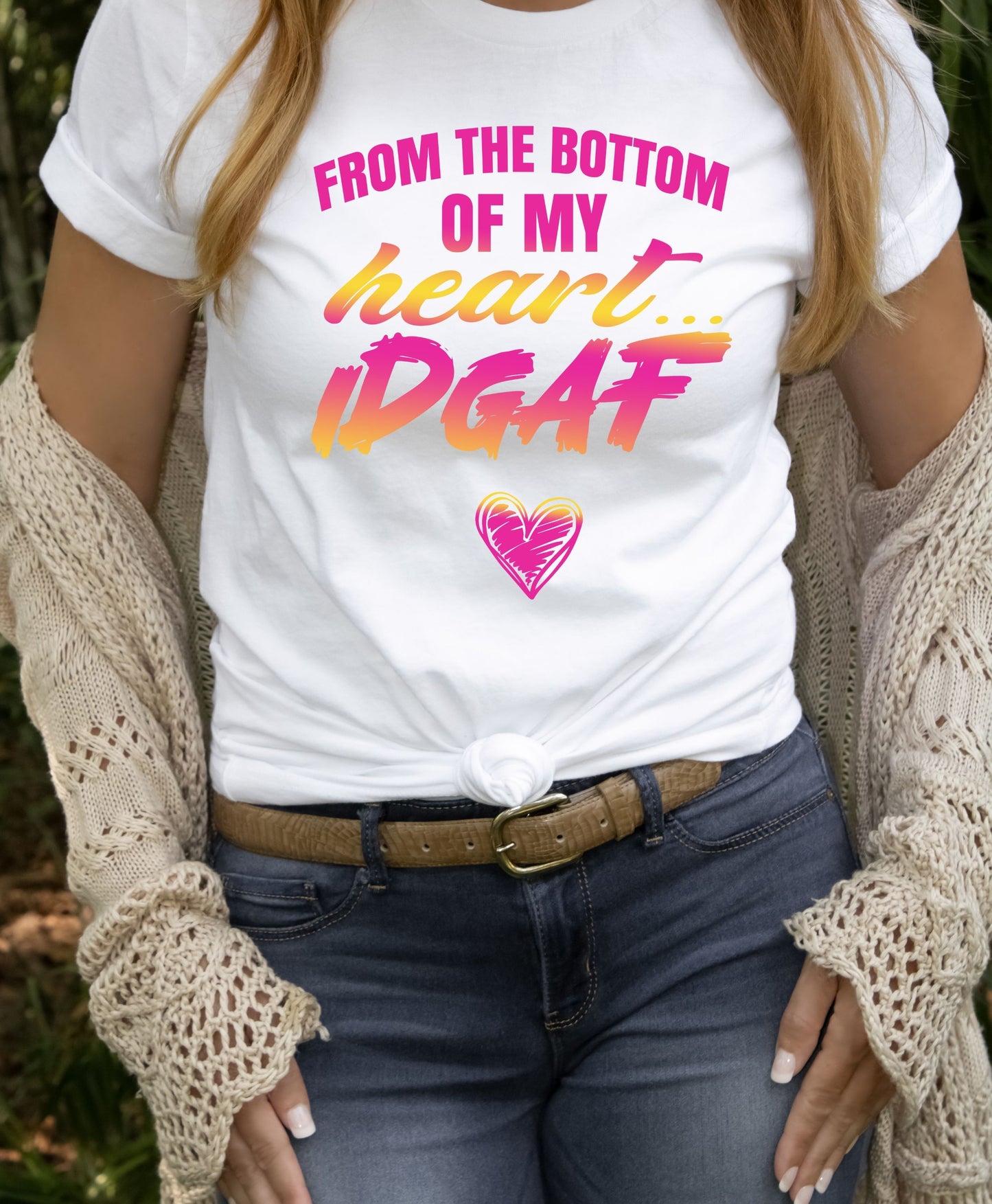 Woman wearing white “From The Bottom Of My Heart… IDGAF” t-shirt with pink and orange gradient lettering, styled with jeans and knit cardigan, holding belt buckle.
