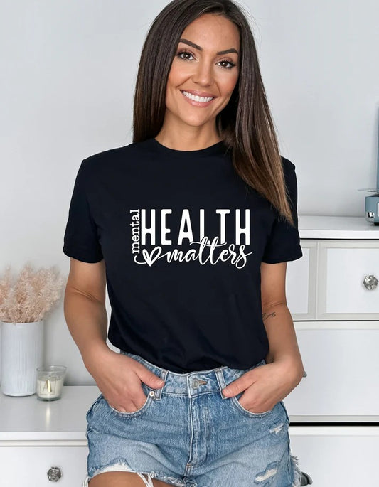 Smiling woman wearing a black Gildan T-shirt with white text reading “Mental Health Matters” and a heart design, paired with denim shorts.