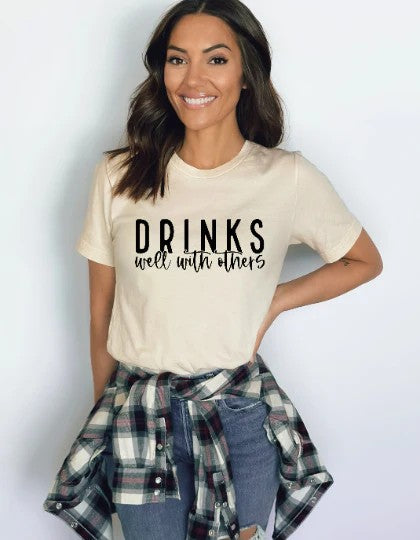 Smiling woman wearing a beige Gildan T-shirt with black text reading “Drinks Well With Others,” styled with ripped jeans and a plaid shirt tied at the waist.