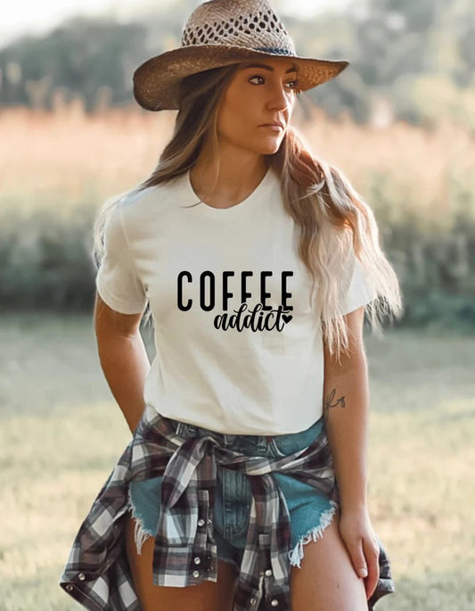 Woman wearing a white Gildan T-shirt with black text reading “Coffee Addict,” styled with denim shorts, a plaid shirt tied at the waist, and a straw hat in an outdoor setting.
