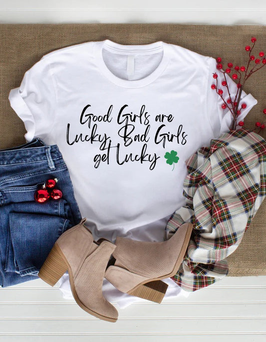 Flat lay of a white Gildan T-shirt with black text reading “Good Girls are Lucky, Bad Girls Get Lucky” and a small green shamrock, styled with jeans, ankle boots, and a plaid scarf for St. Patrick’s Day.