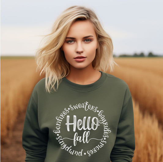 Blonde woman wearing an olive green sweatshirt with the phrase “Hello Fall” surrounded by words like bonfires, sweaters, hayrides, s’mores, and pumpkins in white script font, standing in a golden wheat field.