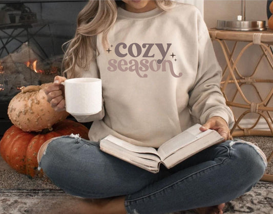 Woman wearing a beige “Cozy Season” sweatshirt with retro text and star accents, sitting by pumpkins, holding coffee and a book