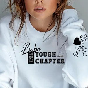 Woman wearing white crewneck sweatshirt with “Babe It’s a Tough Chapter” motivational quote